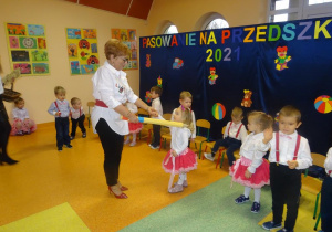 Pani dyrektor Maria Królikowska trzyma w ręku żółtą kredkę, którą przykłada dziewczynce do lewego ramienia.